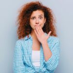 A young woman holds her fingers to her mouth with an expression of shock/uneasiness because she's just heard or used vulgar vocabulary