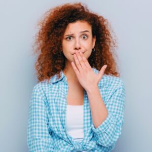 A young woman holds her fingers to her mouth with an expression of shock/uneasiness because she's just heard or used vulgar vocabulary